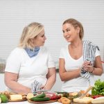 Two women in a kitchen preparing fresh vegetables and cheese with olive oil and spices.